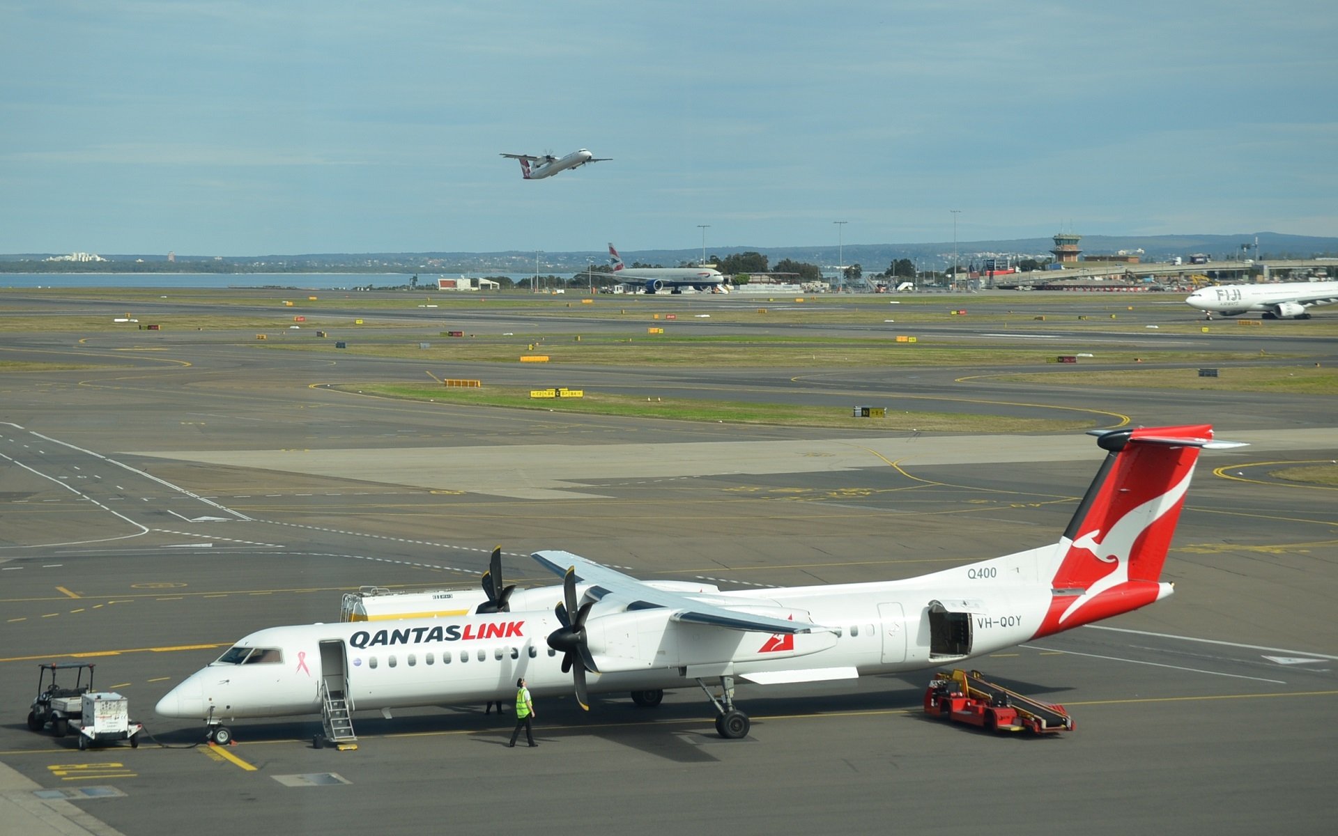 QantasLink Bombardier Dash 8 aircraft parked at Sydney airport, with ground vehicles nearby and another plane taking off in the background.