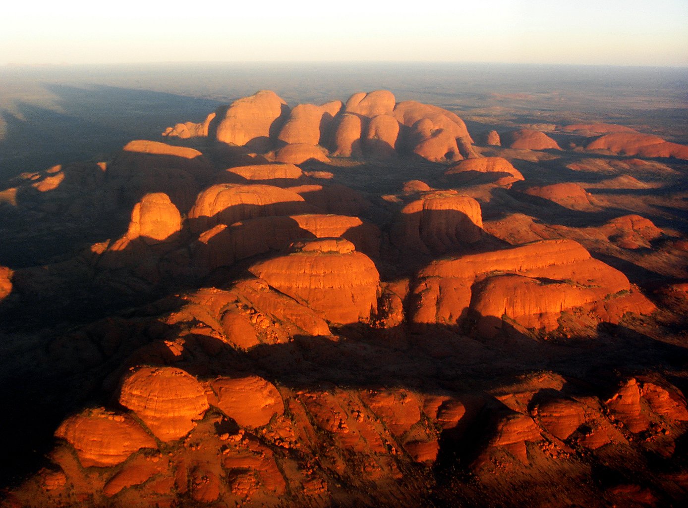 HD desktop wallpaper: aerial view of the Olgas (Kata Tjuta) in Uluru-Kata Tjuta National Park at sunrise, warm red domes casting long shadows across the desert.