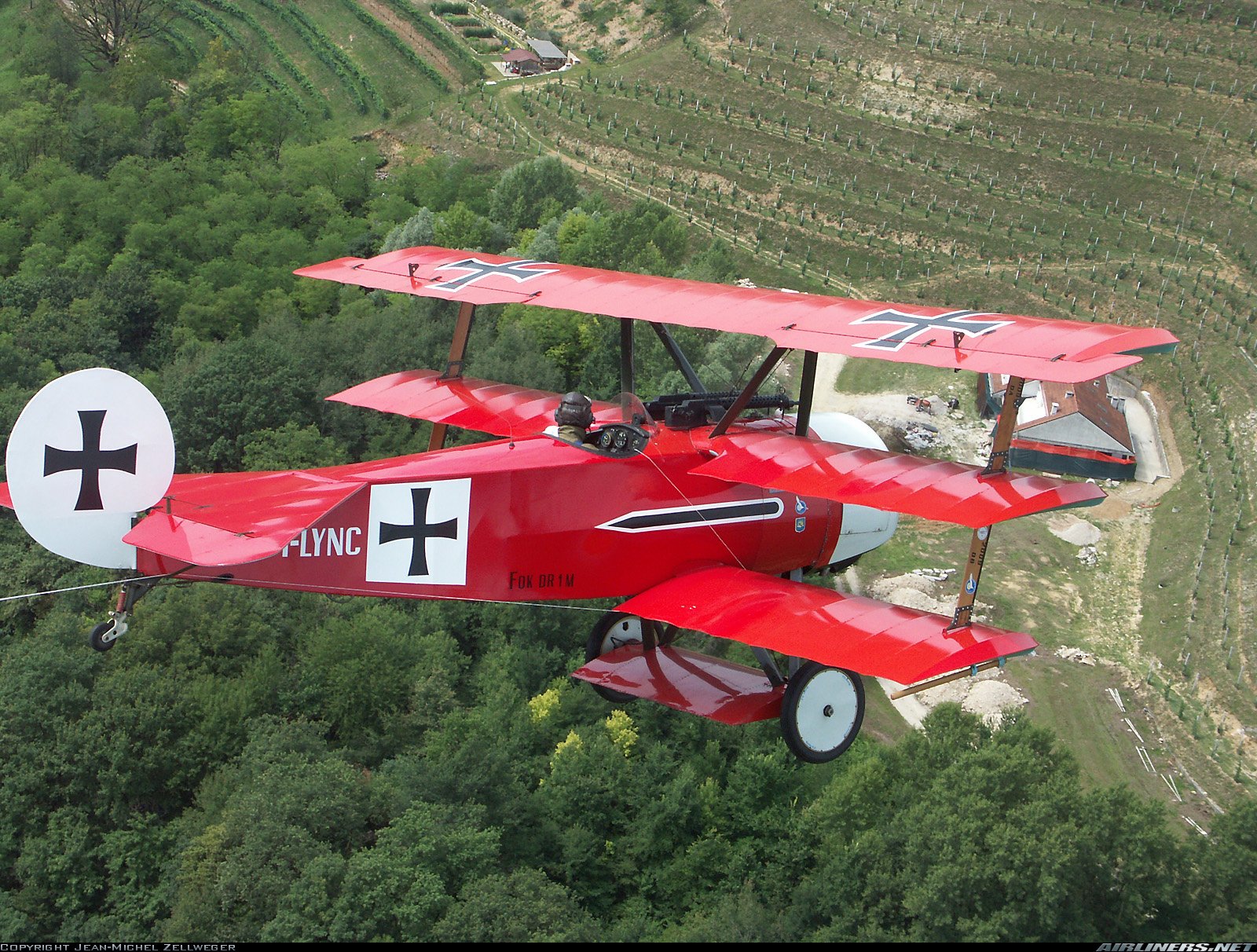 HD desktop wallpaper featuring a red Fokker Dr.I triplane airplane flying over a green forested landscape.