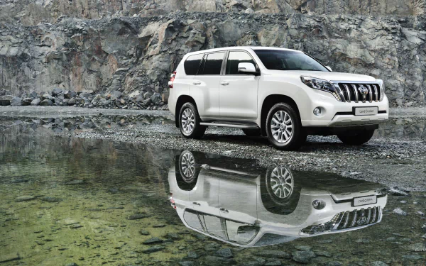HD PC desktop wallpaper featuring a white 2014 Toyota Land Cruiser parked near a rocky water edge, with its reflection clearly visible in the calm water.
