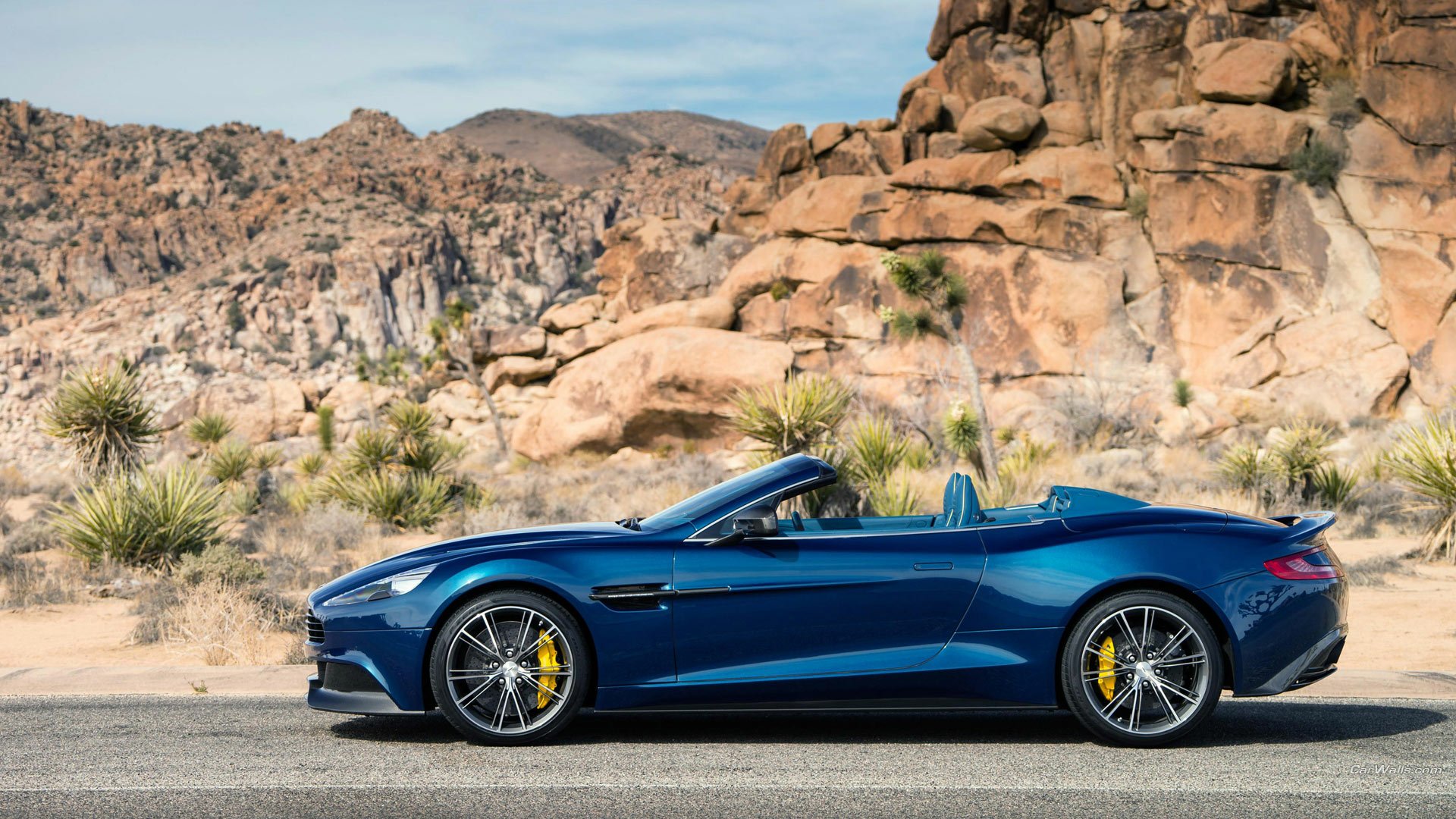Blue 2014 Aston Martin Vanquish Volante convertible vehicle, side profile on a desert road with rocky outcrops — HD PC desktop wallpaper background.