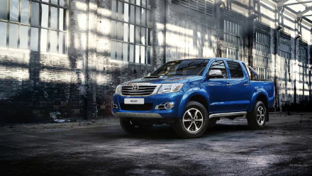 HD desktop wallpaper featuring a blue Toyota Hilux truck parked inside an industrial warehouse with large windows casting light on the vehicle.