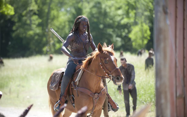 HD wallpaper featuring Michonne from The Walking Dead, portrayed by Danai Gurira, riding a horse in a vibrant, post-apocalyptic landscape with walkers in the background.