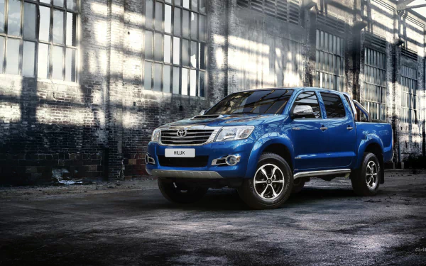 HD desktop wallpaper featuring a blue Toyota Hilux truck parked inside an industrial warehouse with large windows casting light on the vehicle.