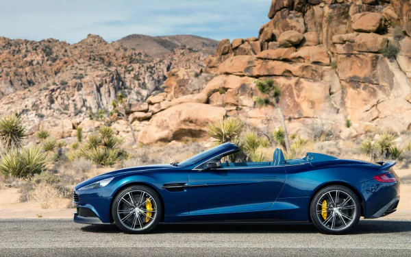 Blue 2014 Aston Martin Vanquish Volante convertible vehicle, side profile on a desert road with rocky outcrops — HD PC desktop wallpaper background.