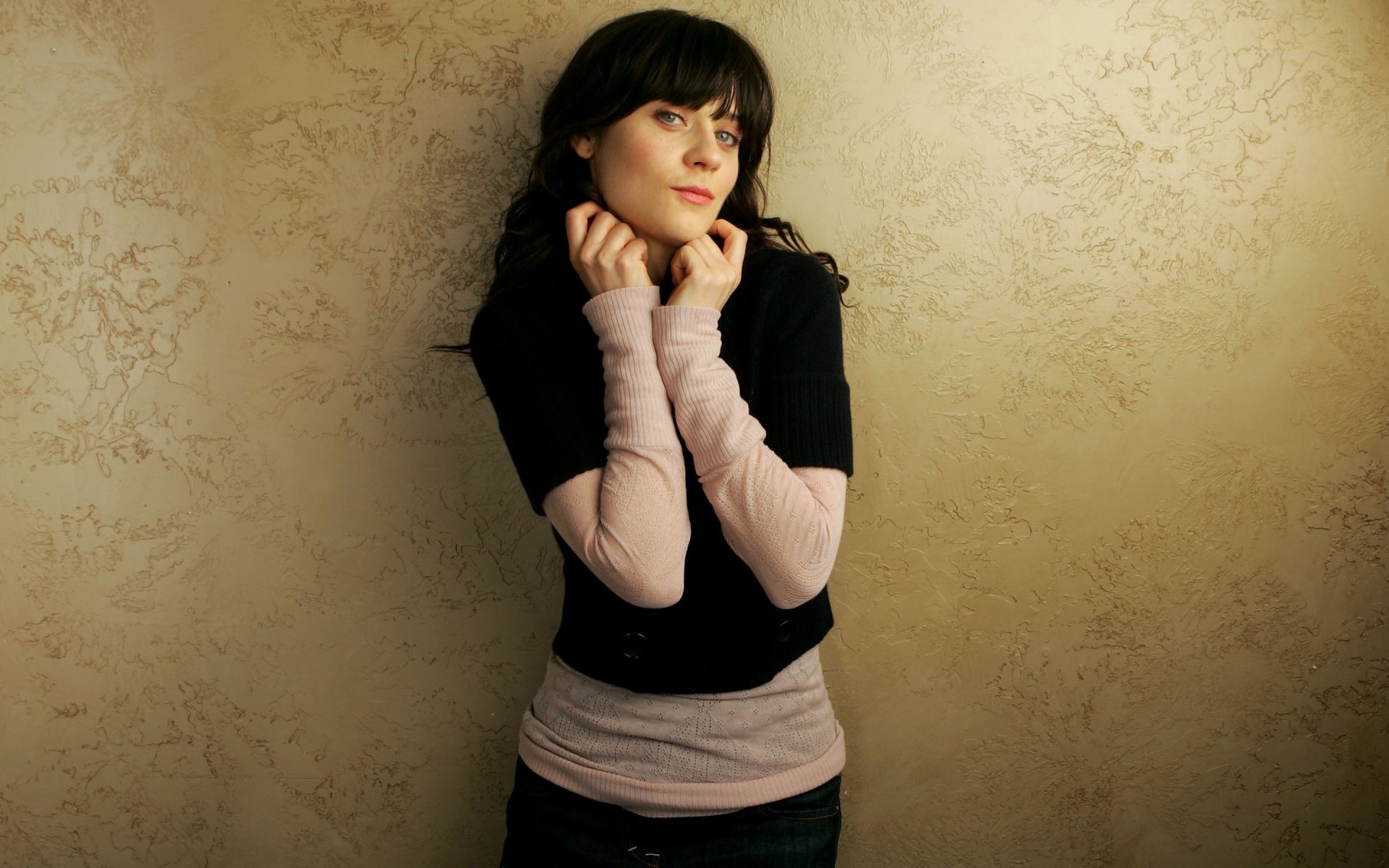 Zooey Deschanel poses thoughtfully against a textured beige wall, showcasing a stylish outfit. This HD image serves as a vibrant desktop wallpaper and background.