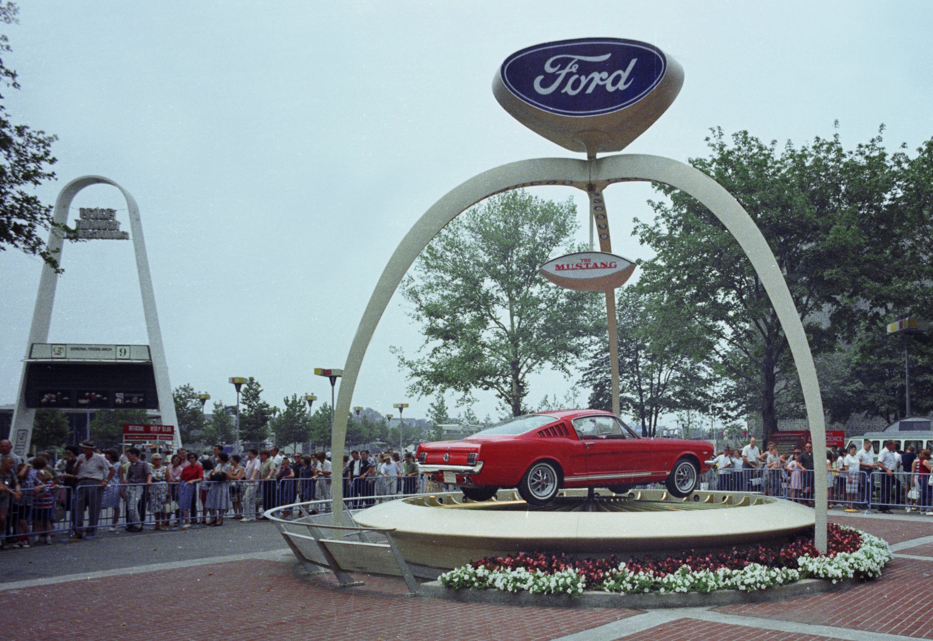 Red Ford Mustang on a raised circular display beneath a Ford arch at an outdoor car show, crowd in the background — 4K Ultra HD PC desktop wallpaper/background.