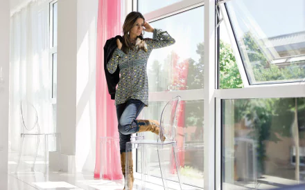 HD PC desktop wallpaper featuring Alessia Ventura, a woman standing by a large window with sunlight streaming in, dressed casually and looking outside contemplatively.