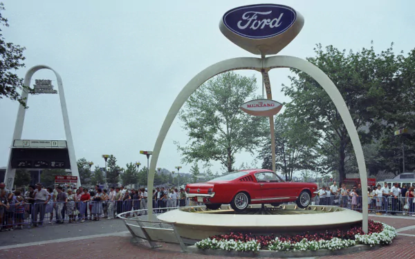 Red Ford Mustang on a raised circular display beneath a Ford arch at an outdoor car show, crowd in the background — 4K Ultra HD PC desktop wallpaper/background.