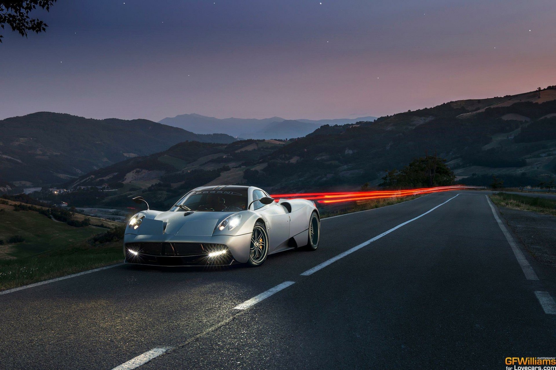 A striking Pagani Huayra positioned on a winding road, capturing motion with light trails against a twilight mountain backdrop. An HD desktop wallpaper that emphasizes automotive elegance.