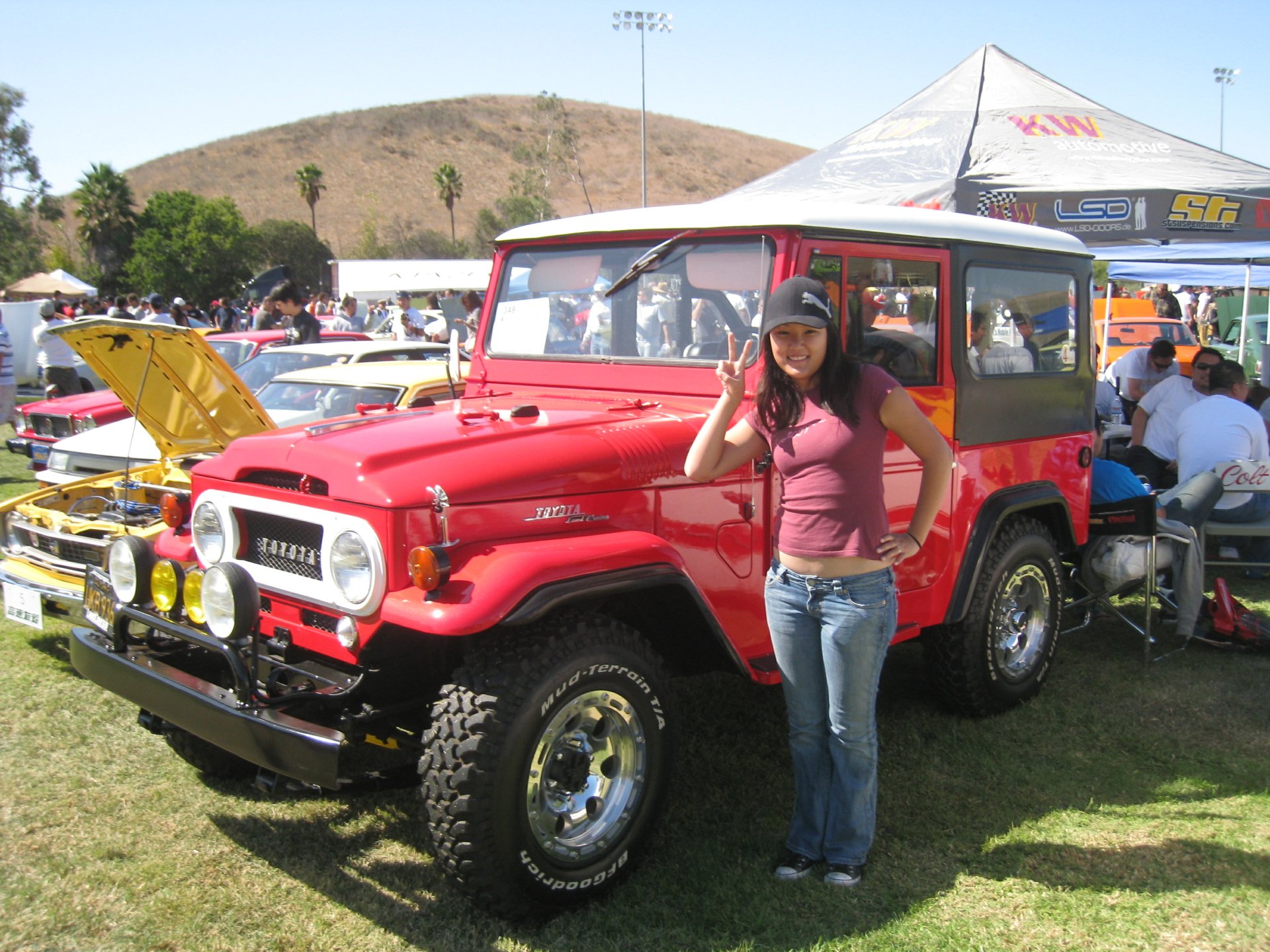 Red Toyota Land Cruiser at a car show with a woman posing beside it, vibrant 2K Quad HD PC desktop wallpaper and background