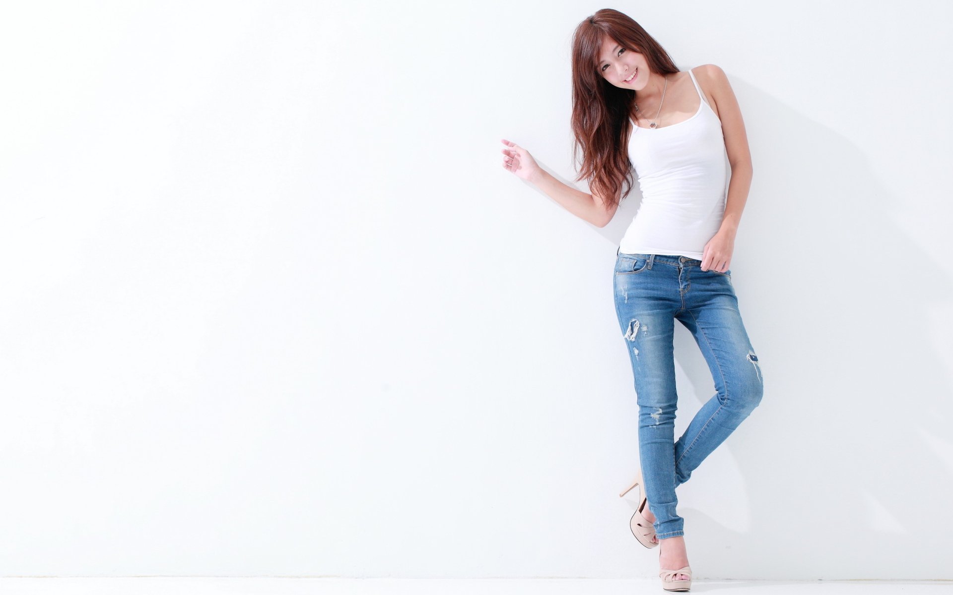 HD wallpaper of an Asian woman wearing a white tank top and blue jeans, leaning against a white wall with a relaxed pose.