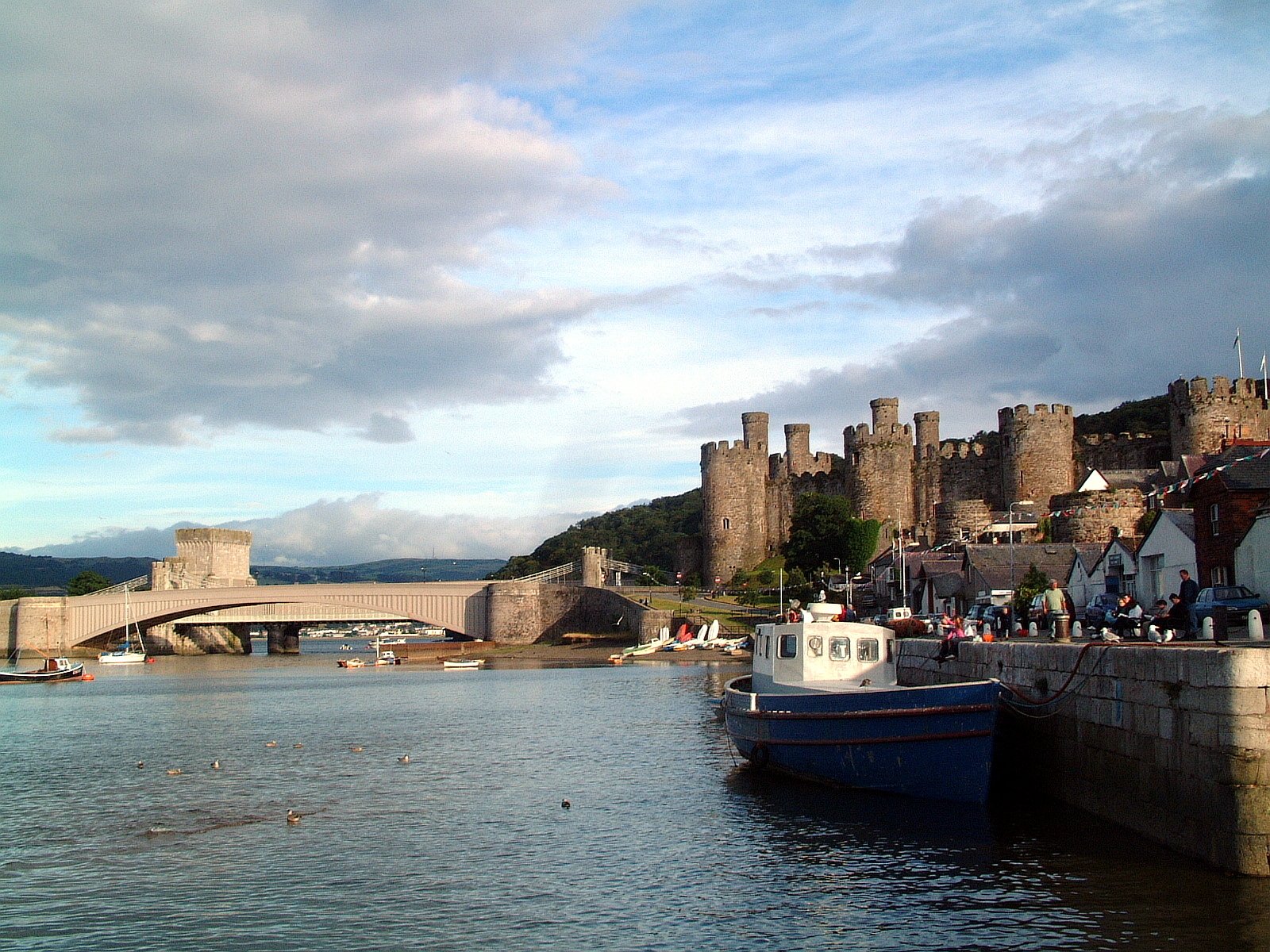 Man-made Conwy Castle and town walls overlooking the harbor with boats and a bridge under a cloudy sky — HD PC desktop wallpaper/background.