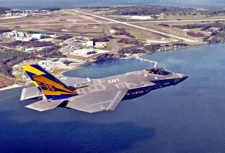 An HD desktop wallpaper featuring a Lockheed Martin F-35 Lightning II military jet flying over a scenic landscape with water and an airfield in the background.