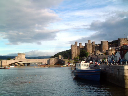 Man-made Conwy Castle and town walls overlooking the harbor with boats and a bridge under a cloudy sky — HD PC desktop wallpaper/background.
