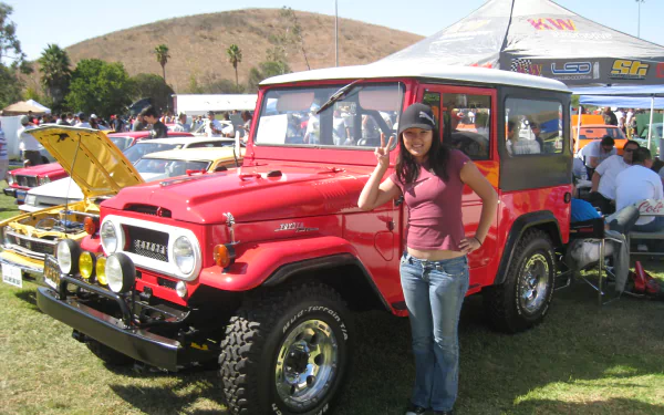 Red Toyota Land Cruiser at a car show with a woman posing beside it, vibrant 2K Quad HD PC desktop wallpaper and background
