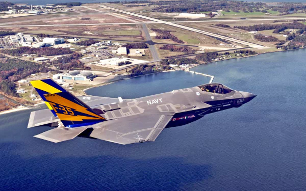 An HD desktop wallpaper featuring a Lockheed Martin F-35 Lightning II military jet flying over a scenic landscape with water and an airfield in the background.