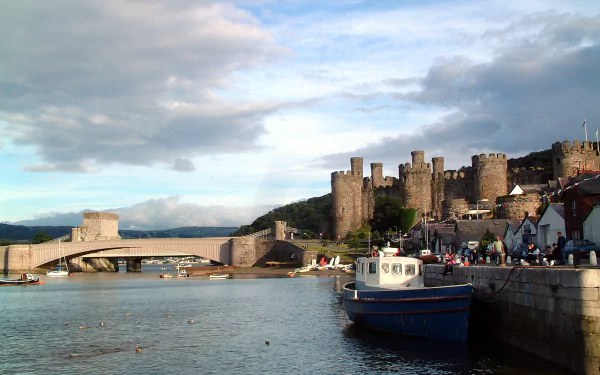 Man-made Conwy Castle and town walls overlooking the harbor with boats and a bridge under a cloudy sky — HD PC desktop wallpaper/background.