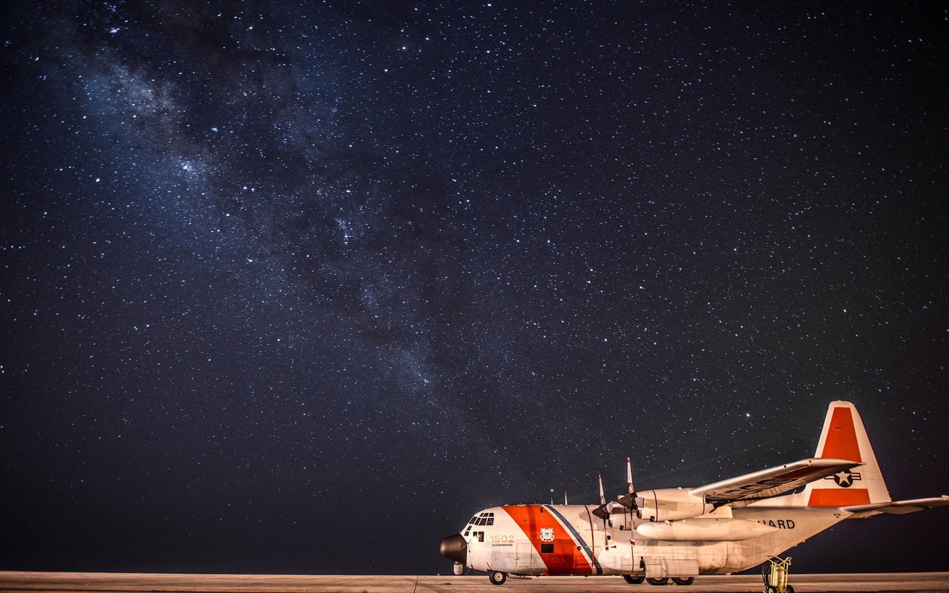HD desktop wallpaper of a Lockheed C-130 Hercules military aircraft parked under a starry night sky with the Milky Way visible.