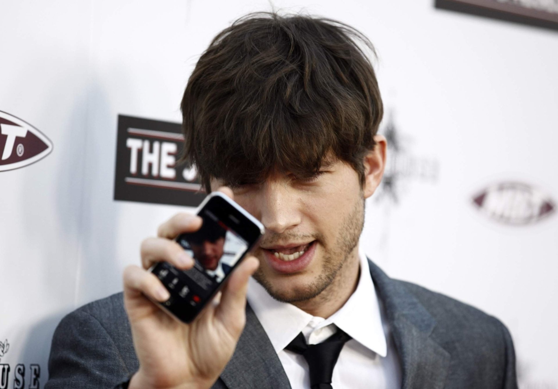 Close-up of a man in a suit holding a phone toward the camera against a branded event backdrop — HD desktop wallpaper.