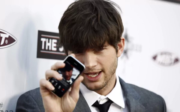 Close-up of a man in a suit holding a phone toward the camera against a branded event backdrop — HD desktop wallpaper.