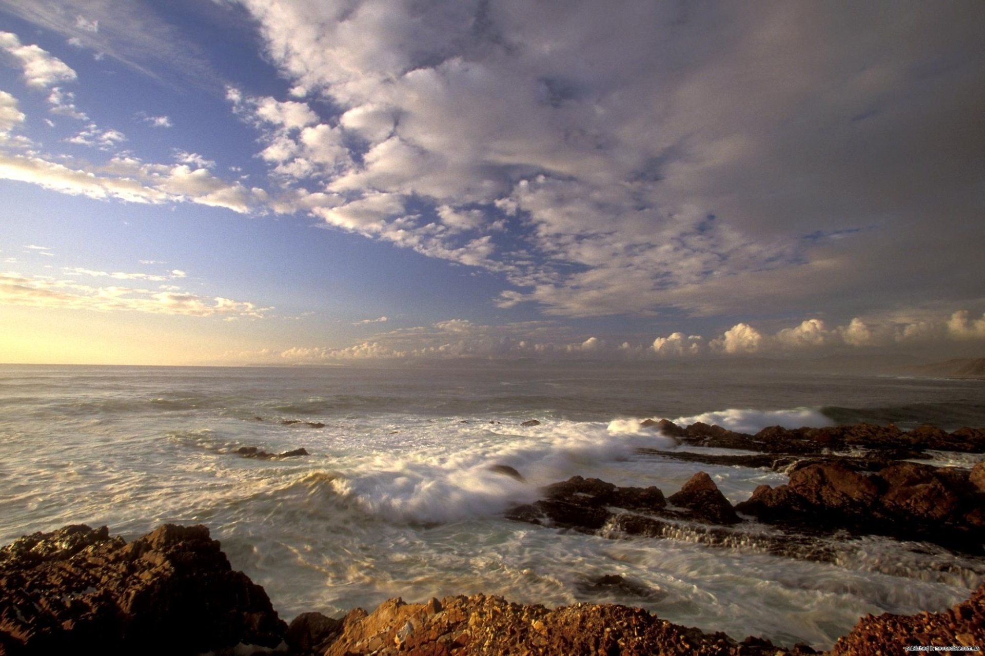HD desktop wallpaper featuring a dramatic ocean landscape with rocky shore, waves, and a sky filled with scattered clouds at sunrise or sunset.