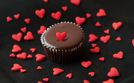 A chocolate cupcake with a red heart on top is surrounded by scattered red heart sprinkles on a black background. This HD image makes a delightful desktop wallpaper celebrating love and delicious food.