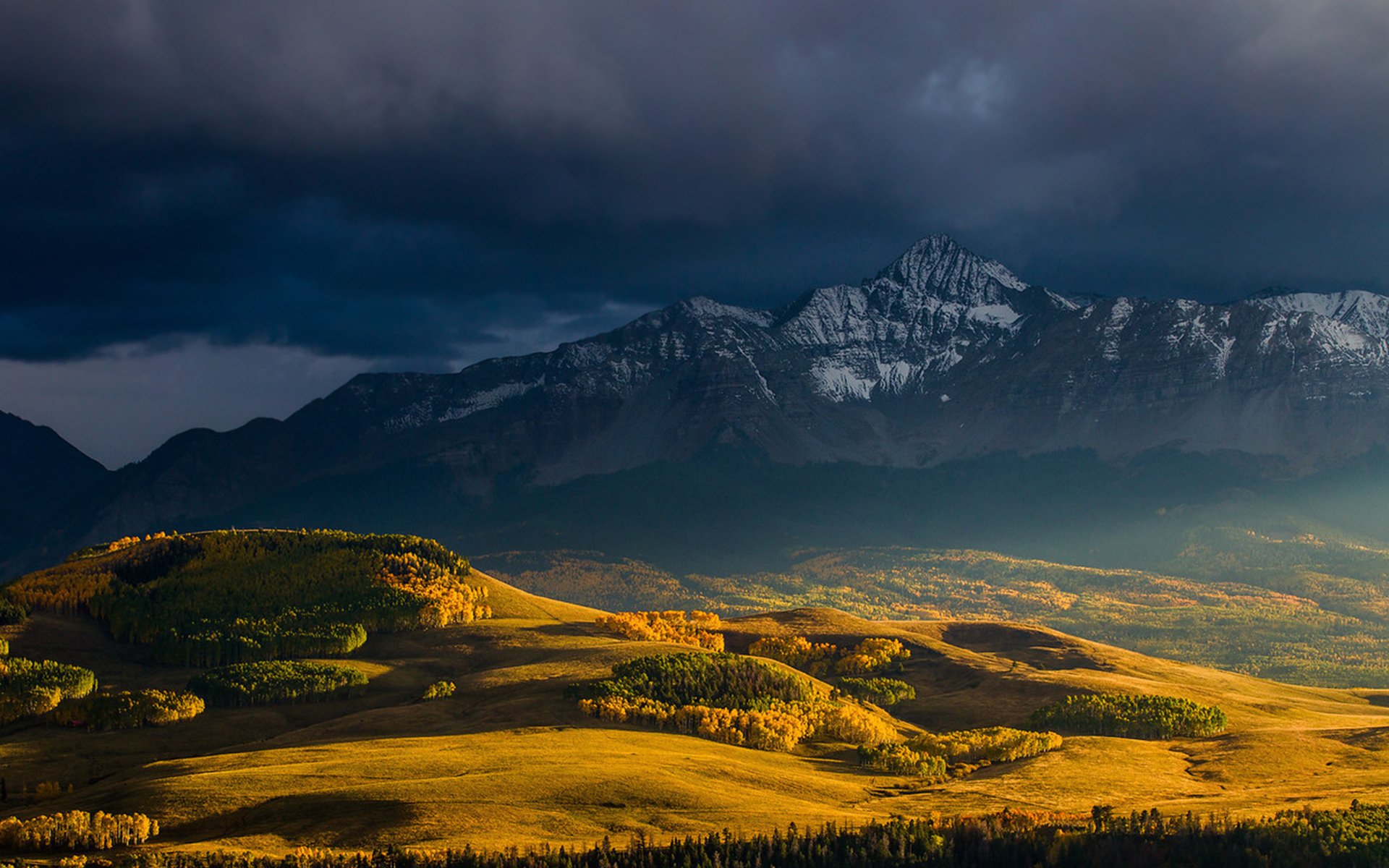 HD PC desktop wallpaper showcasing vibrant fall foliage on rolling hills under a dramatic sky with towering mountains in the background.