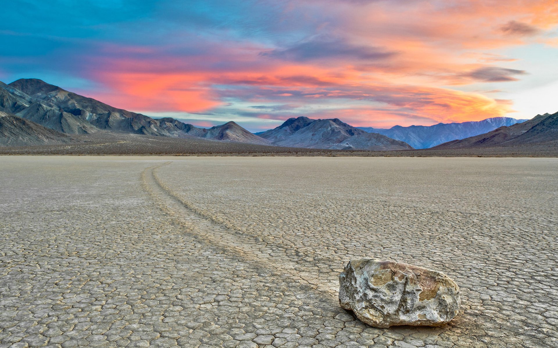 Nature desert HD PC desktop wallpaper: a lone rock leaves a winding trail across a cracked dry playa toward distant mountains beneath a colorful sunset.