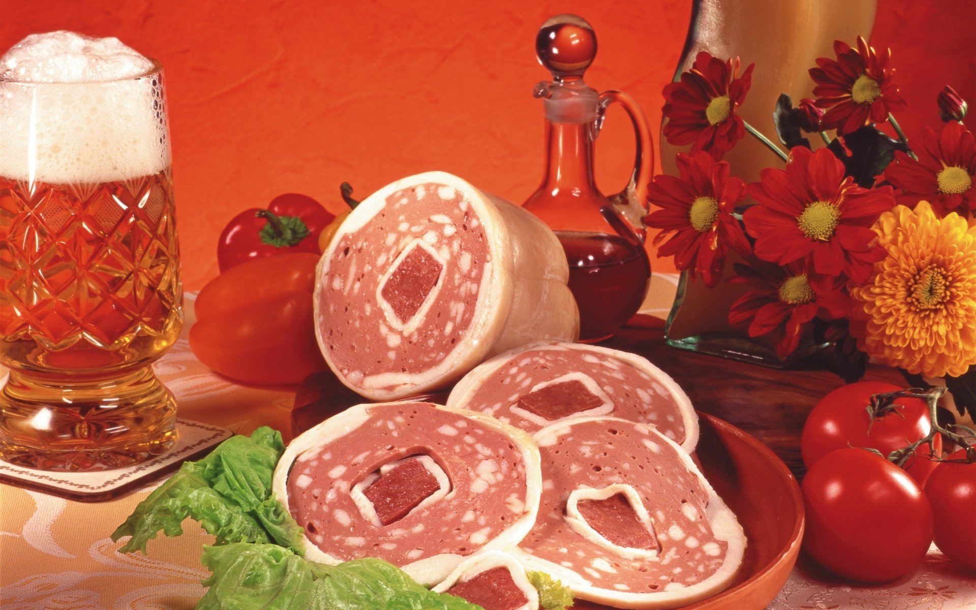 HD PC desktop wallpaper featuring sliced cured meat on a plate, accompanied by fresh vegetables, a glass of beer, a decanter, and vibrant flowers against a warm orange background.