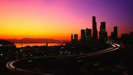 HD PC desktop wallpaper of Seattle skyline at sunset, orange-purple sky over downtown skyscrapers and a curving illuminated highway, man-made cityscape.