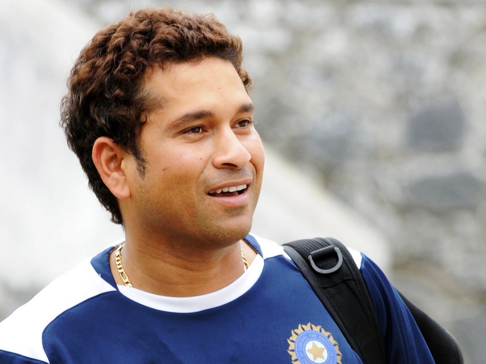 HD desktop wallpaper featuring a close-up of a cricket player smiling, wearing a navy blue sports jersey with a bag strap over his shoulder.