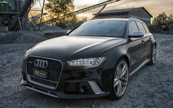 HD desktop wallpaper featuring a sleek black Audi RS6 parked on a gravel surface with industrial structures in the background at sunset.