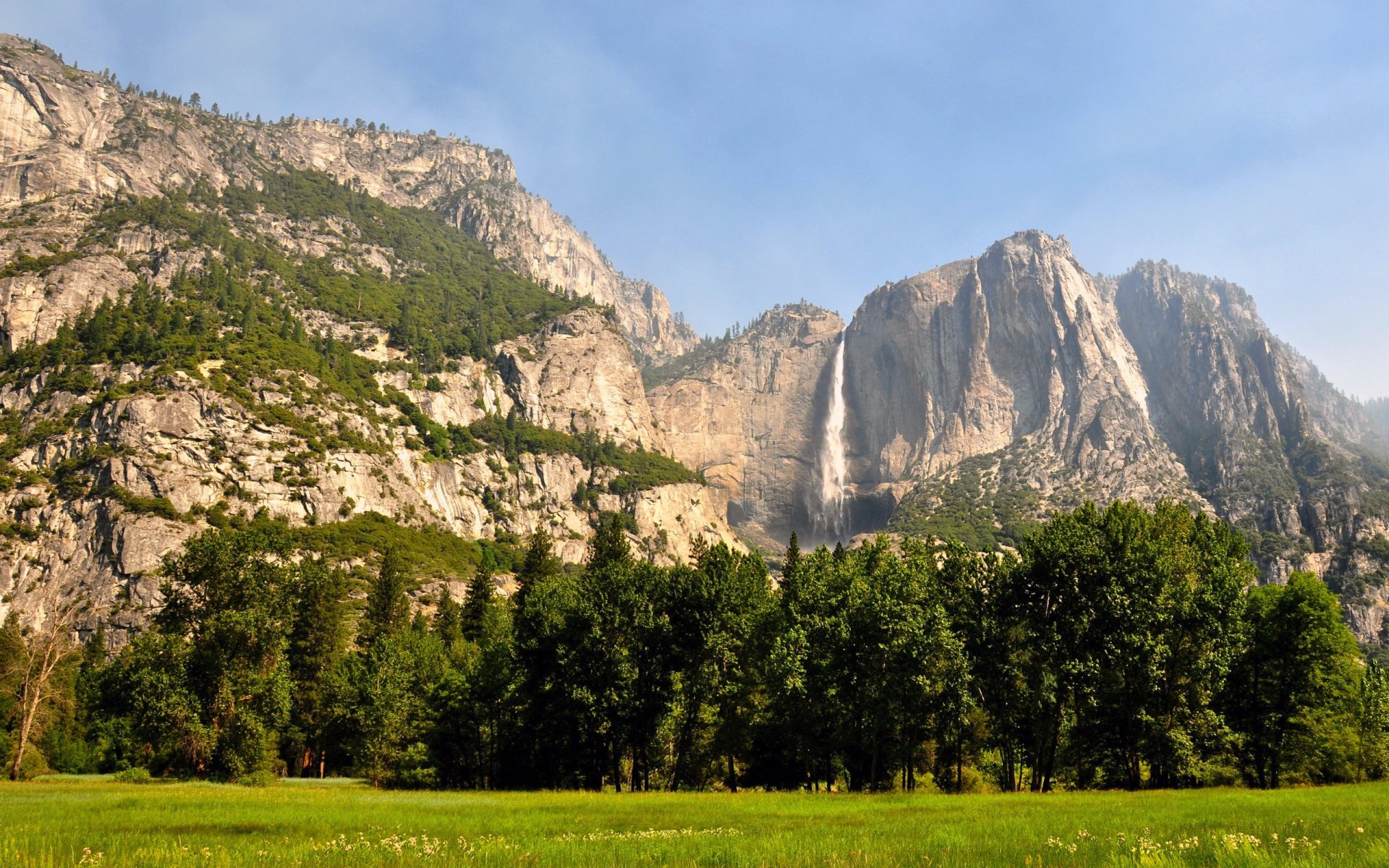 HD landscape wallpaper showing a mountain cliff with a waterfall, dense forest, tall trees, and a wide grassy area under a clear blue sky.