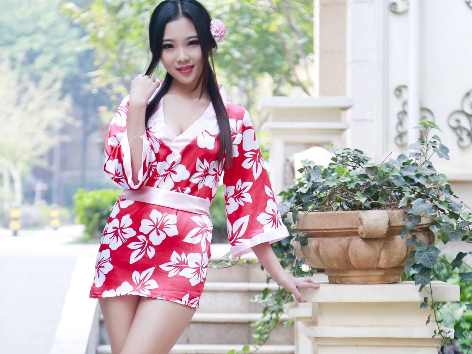 Asian woman in a red floral dress poses outdoors beside a potted ivy plant. This HD desktop wallpaper captures an elegant, oriental aesthetic.