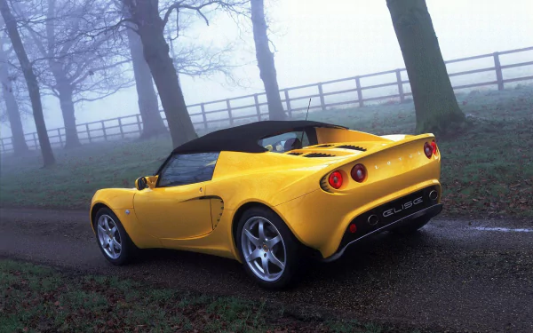 HD desktop wallpaper featuring a yellow Lotus Elise sports car parked on a foggy road with bare trees and a wooden fence in the background.