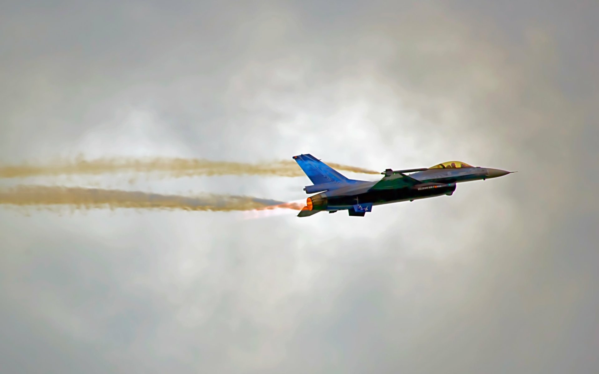 HD desktop wallpaper featuring a General Dynamics F-16 Fighting Falcon military aircraft soaring through a cloudy sky with afterburners engaged.