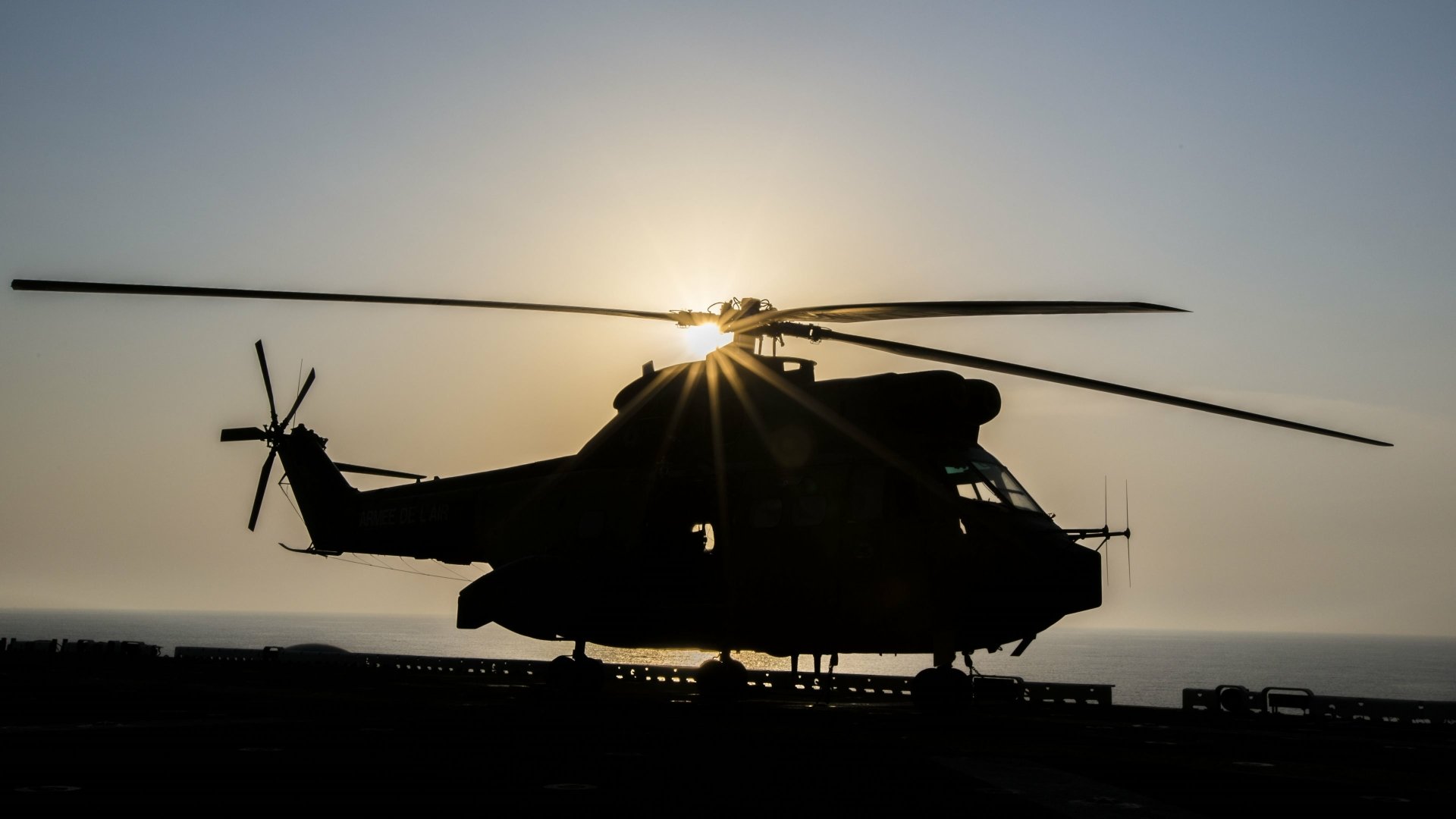 Silhouetted military Aérospatiale SA 330 Puma helicopter on a ship deck at sunset, sunburst through the rotors — 4K Ultra HD PC desktop wallpaper and background.
