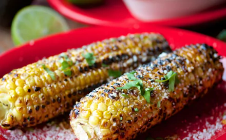 Close-up of two grilled corn cobs topped with cheese and cilantro on a red plate, surrounded by fresh lime, set against a colorful background. HD desktop wallpaper featuring vibrant food.