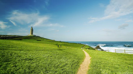 A scenic view of a green field leading to a lighthouse, with waves crashing nearby under a bright blue sky. A tranquil nature setting captured in stunning 4K Ultra HD.
