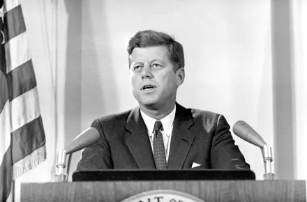 Black and white HD desktop wallpaper of John F. Kennedy delivering a speech, with an American flag in the background.