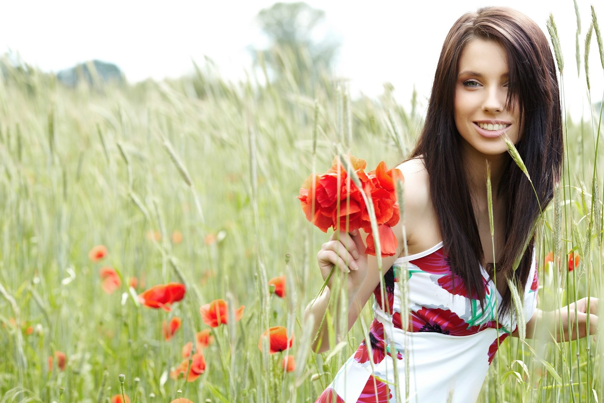 A woman stands in a lush field of tall grass and red poppies, smiling brightly while holding a bouquet of flowers, capturing a vibrant and cheerful mood. HD PC desktop wallpaper.