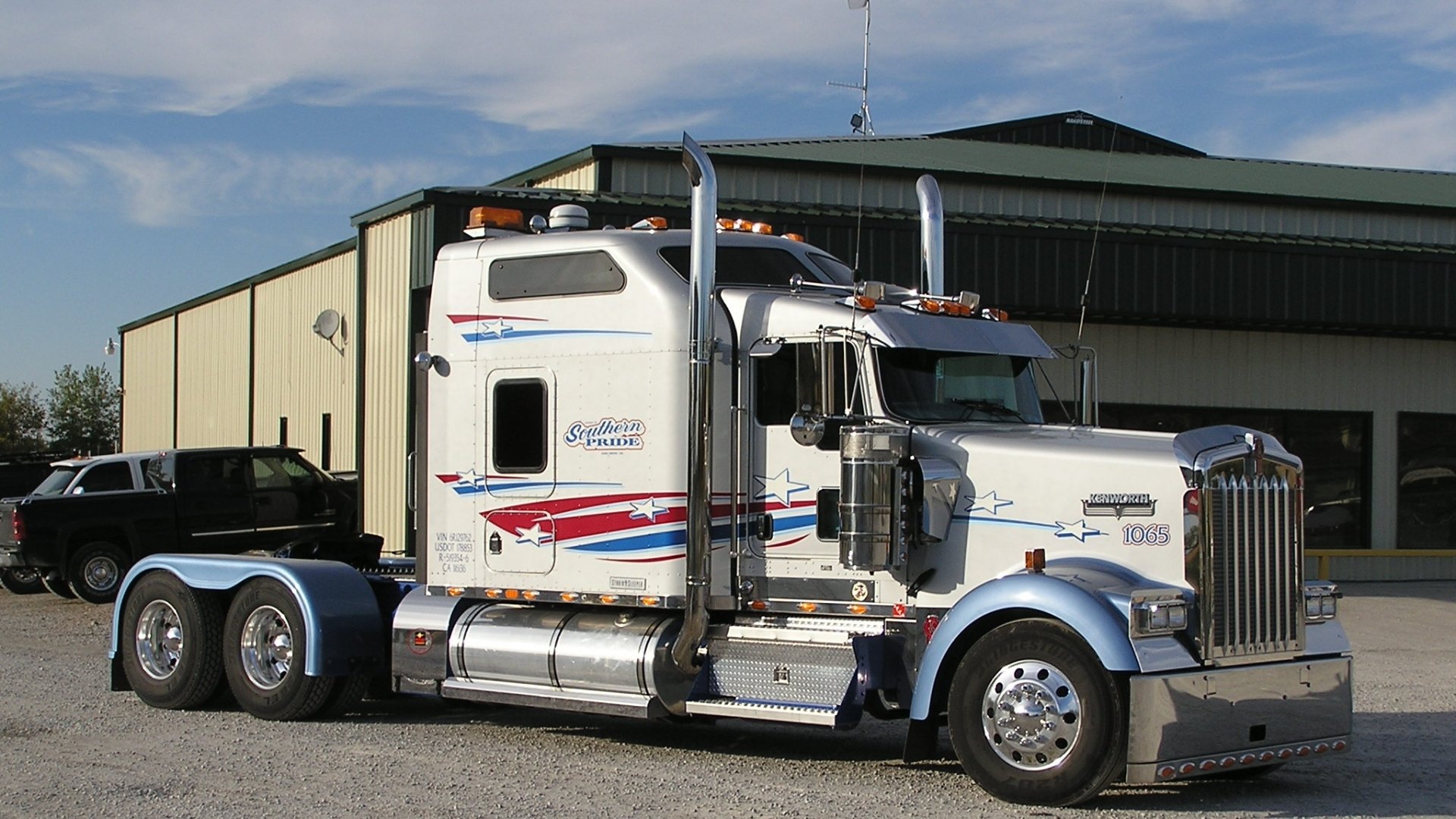 HD desktop wallpaper featuring a white Kenworth truck parked in front of a large industrial building under a partly cloudy sky.