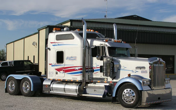 HD desktop wallpaper featuring a white Kenworth truck parked in front of a large industrial building under a partly cloudy sky.