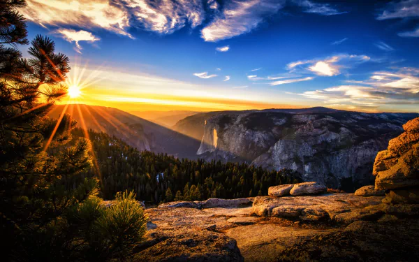 A stunning HD desktop wallpaper featuring a vibrant sunrise over a mountainous landscape, with rays of sunlight illuminating the sky and a forested valley below.