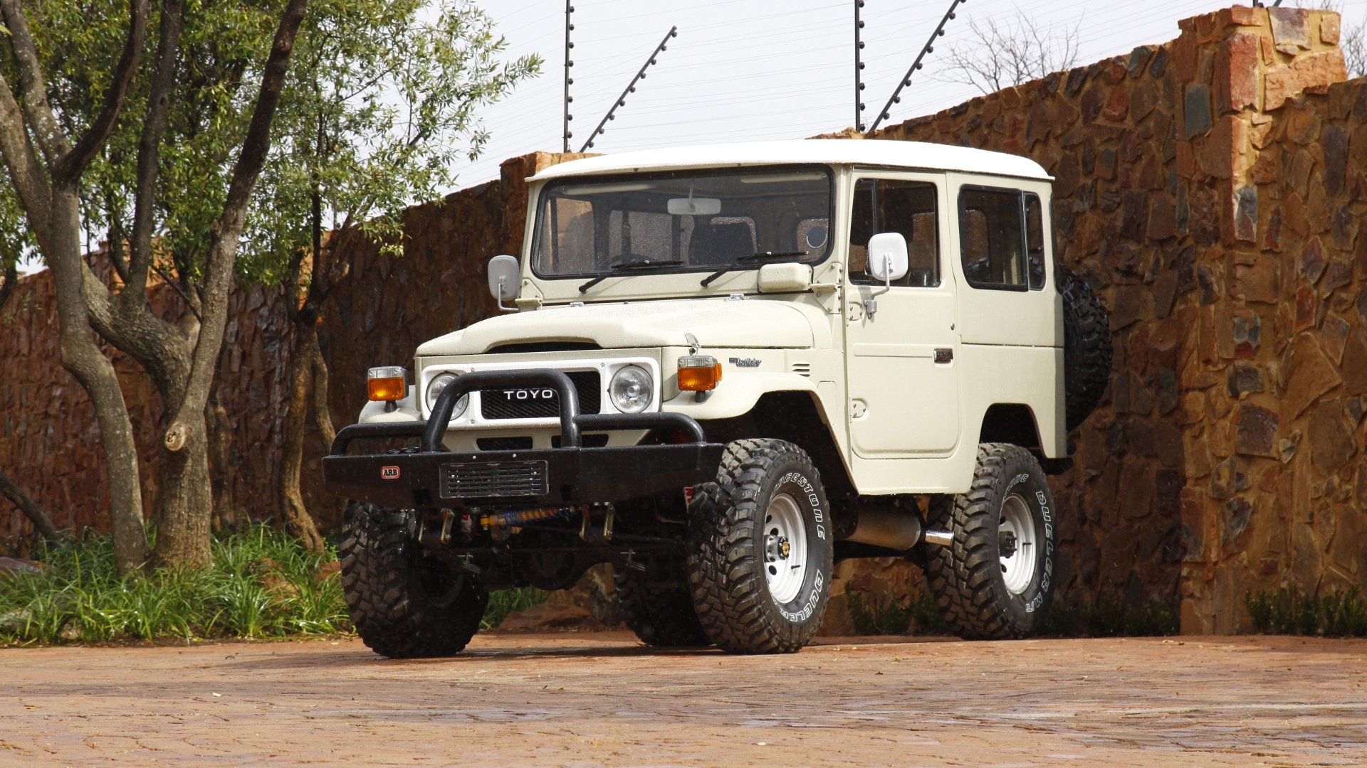8K Ultra HD PC desktop wallpaper/background: classic white Toyota off-road vehicle parked on a stone driveway beside a rustic wall and trees.
