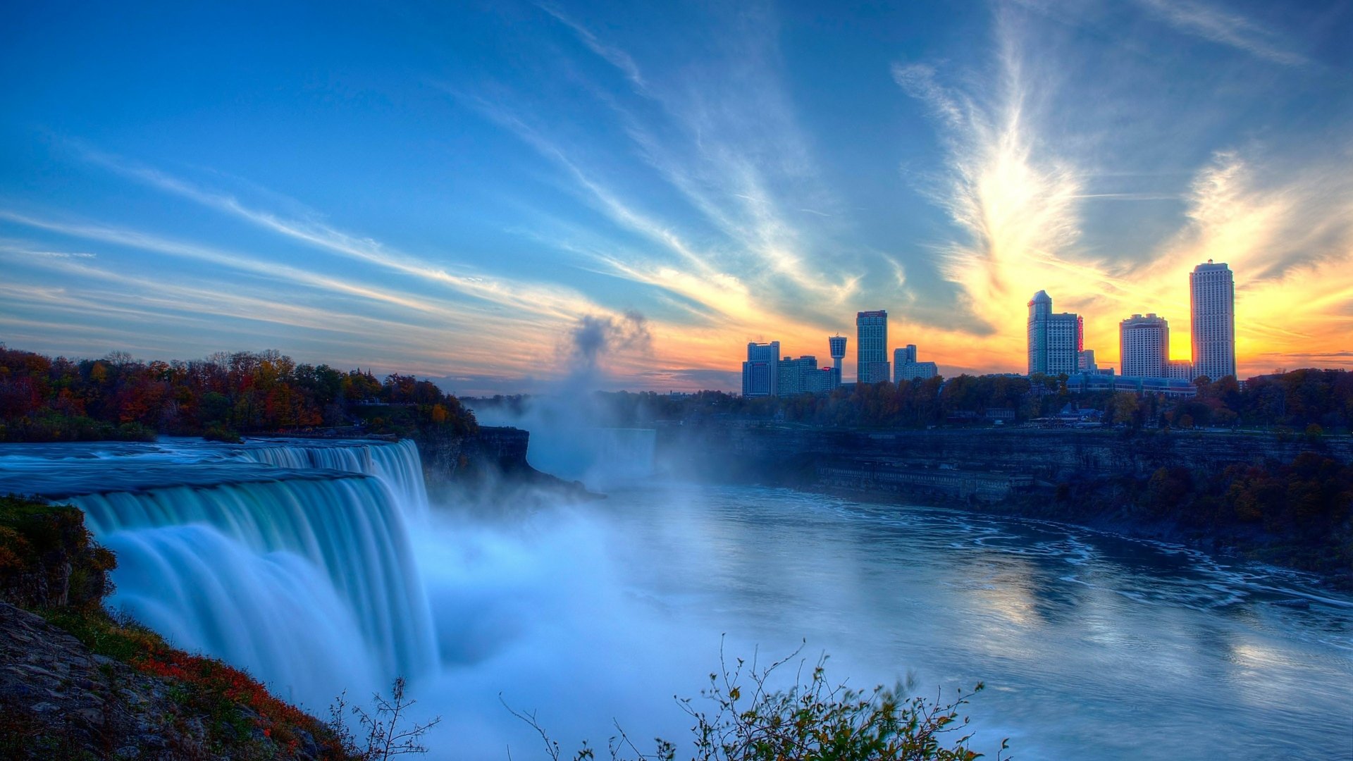 HD desktop wallpaper featuring the majestic Niagara Falls, with a backdrop of a vibrant sunrise and a distant city skyline. Lush greenery frames the cascading waterfalls.
