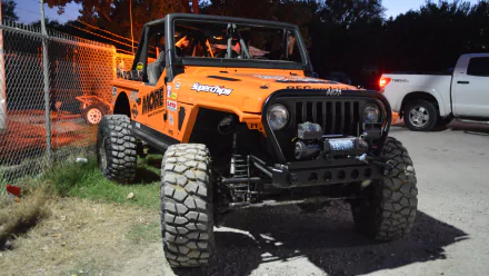 HD desktop wallpaper featuring an orange Jeep with large off-road tires, parked near a fence and another vehicle in a nighttime setting.