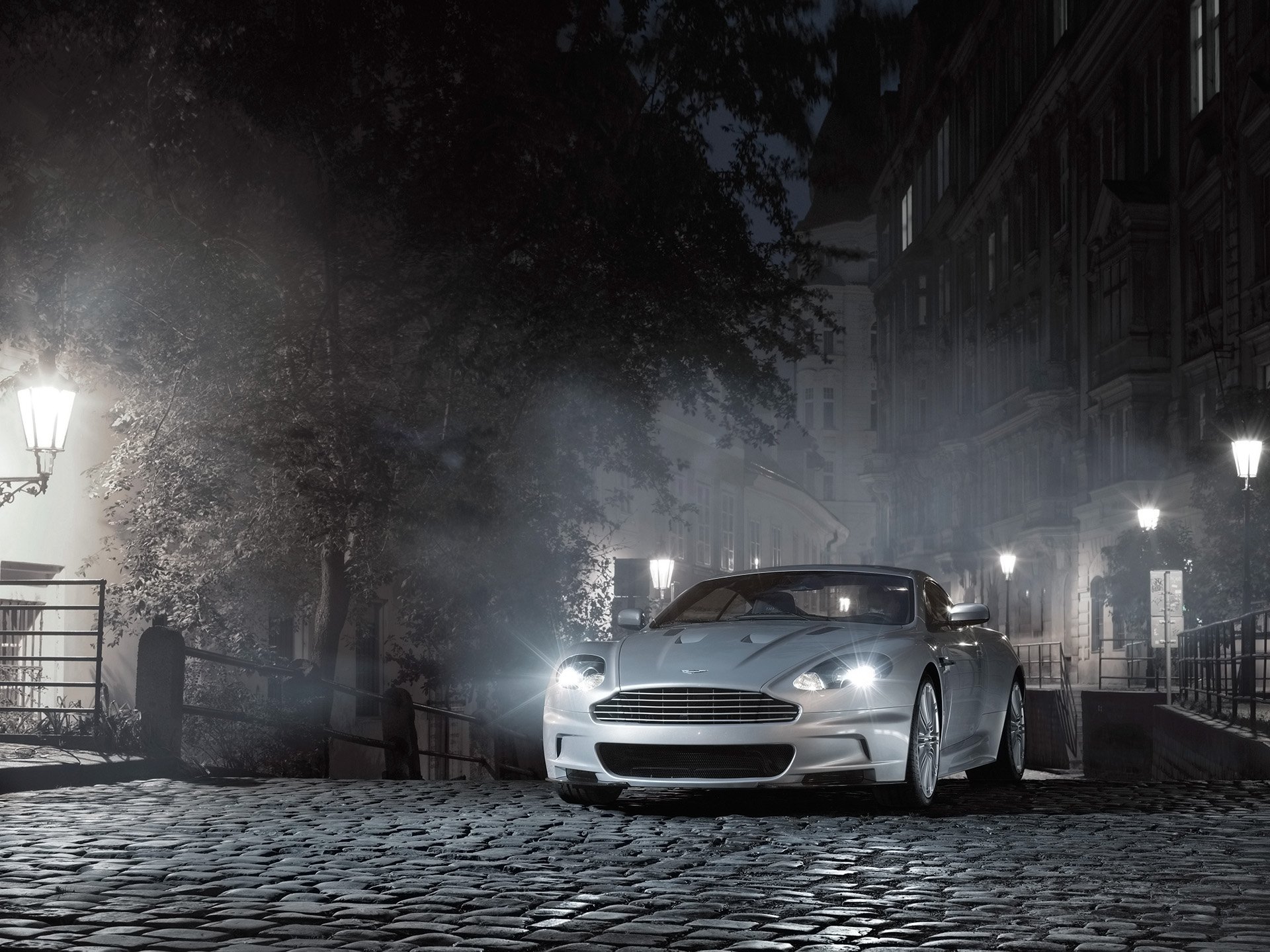 A sleek Aston Martin DBS parked on a cobblestone street at night, illuminated by soft street lamps in an atmospheric, foggy urban setting.