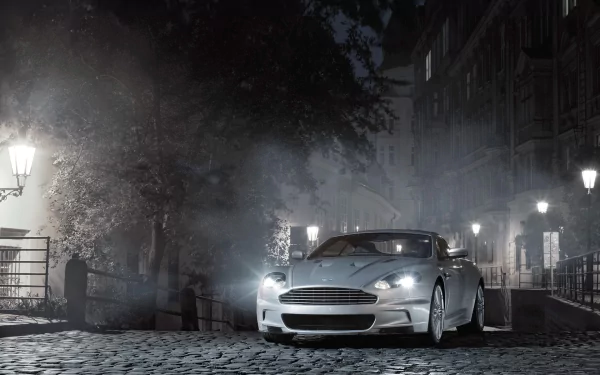 A sleek Aston Martin DBS parked on a cobblestone street at night, illuminated by soft street lamps in an atmospheric, foggy urban setting.
